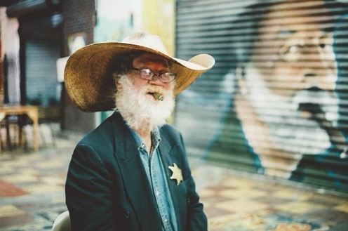 Elderly man in cowboy hat and sheriff's badge smokes cigar outdoors, wearing glasses, with a blurred portrait in the background.