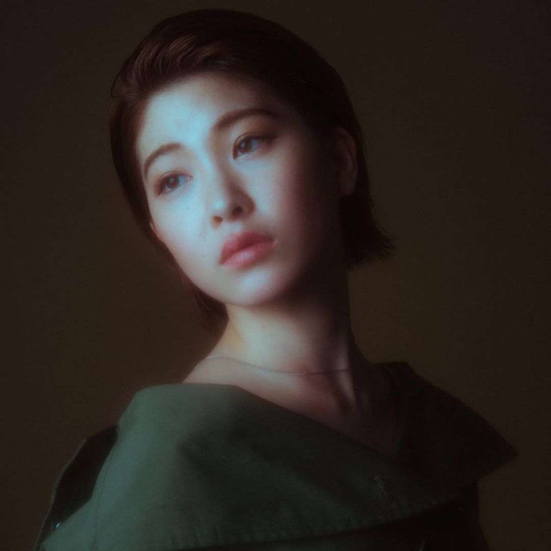 Close-up portrait of a young Asian woman with short brown hair looking away wearing a green collared shirt against a blurred background.