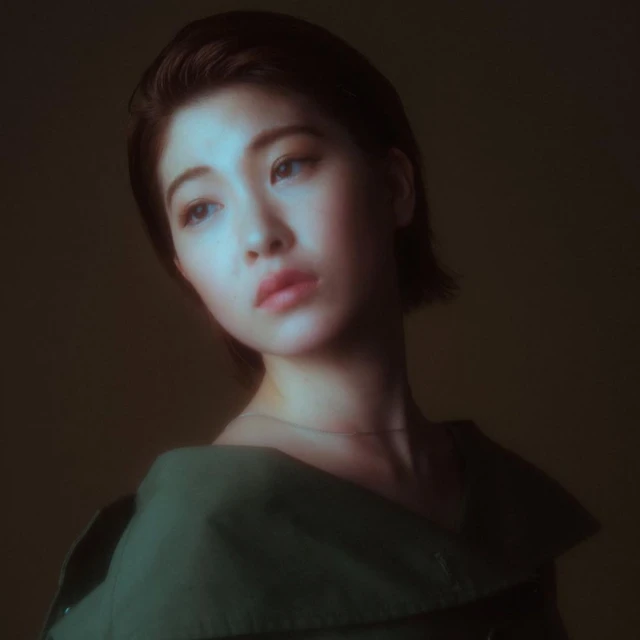 Close-up portrait of a young Asian woman with short brown hair looking away wearing a green collared shirt against a blurred background.