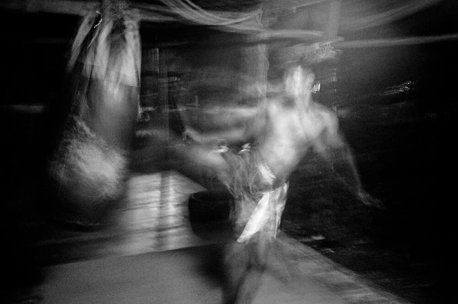 Black-and-white blurred action shot of an adult male kicking a punching bag in an indoor space with subdued lighting and motion blur.