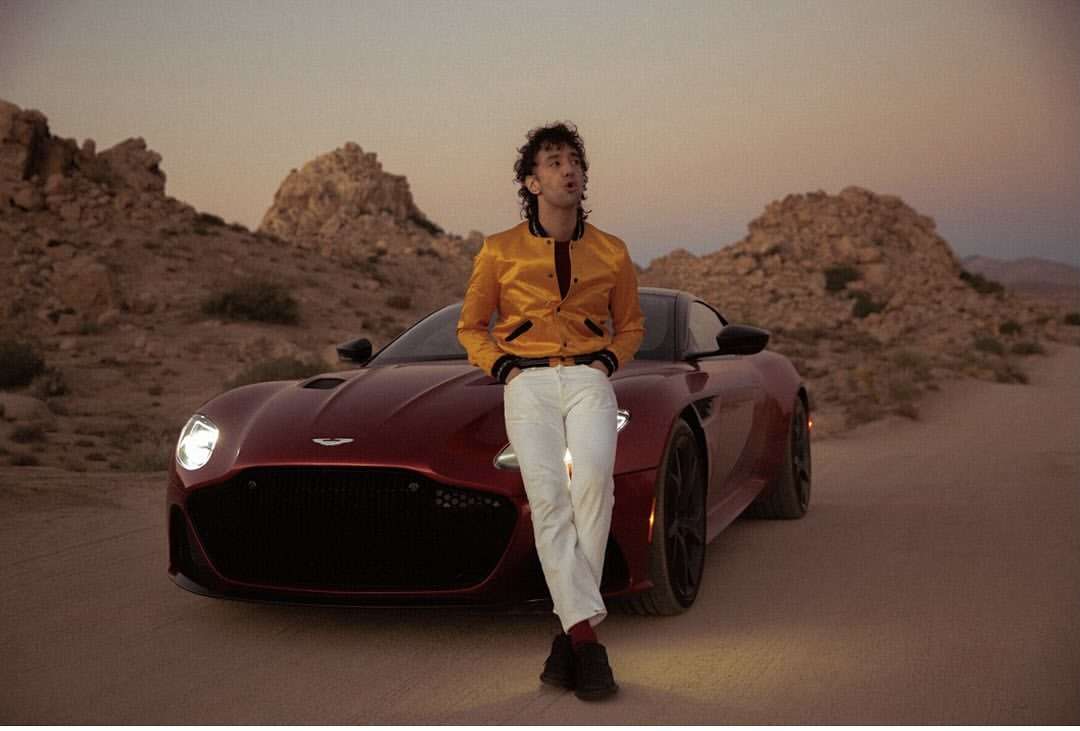 Man in yellow jacket and white pants leans against a red Aston Martin in a desert landscape at sunset, showcasing automotive fashion.