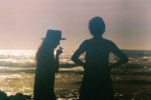 Silhouetted figures on a beach at sunset, one smoking, with gentle waves and soft, dreamy lighting creating a tranquil seascape.