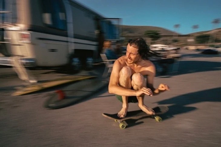 Adult male with dreadlocks crouches on a skateboard on a paved lot. Motorhome and surfboards are in the background on a clear day.