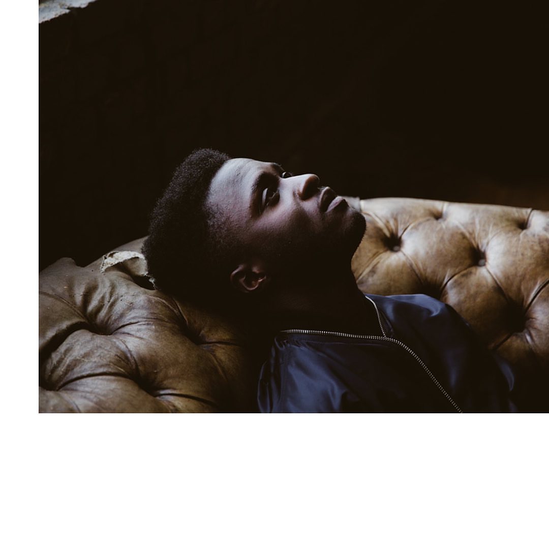 Portrait of a young Black man lying on a brown leather couch, looking up with a serious expression in a dimly lit indoor space.