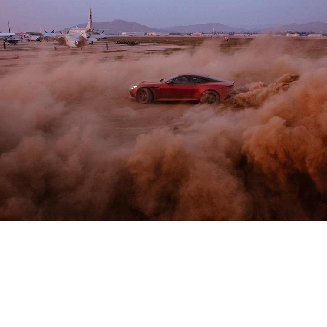A red sports car kicks up dust on an airfield, surrounded by a cloud of brown smoke, with parked airplanes in the background.