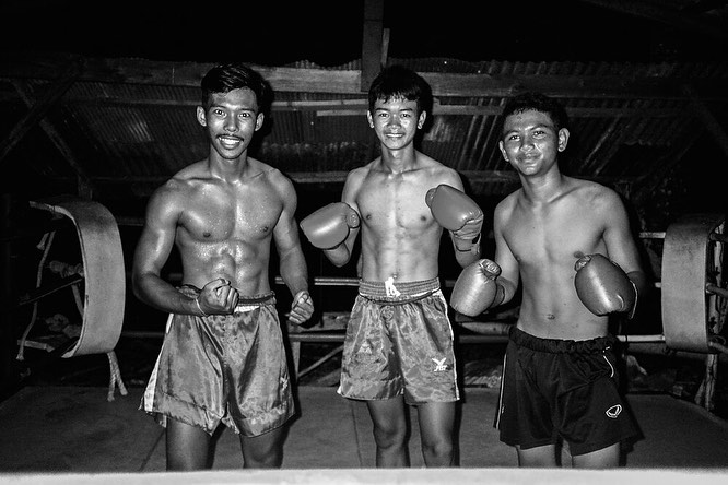 Three shirtless Asian male boxers pose in a boxing ring, wearing shorts and gloves, looking at camera in a monochrome image.
