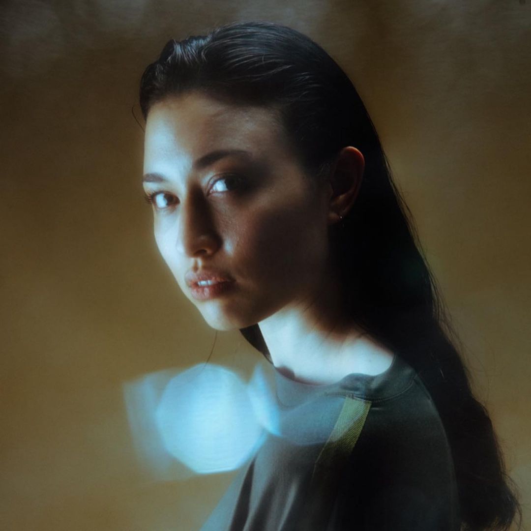 Close-up portrait of a young woman with long dark hair, wearing a dark top, against a tan background with blurred light effects.