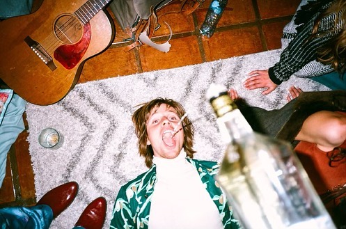 Overhead shot of a young adult man lying on a rug with a guitar, bottle, and other people around him, in a casual indoor setting.