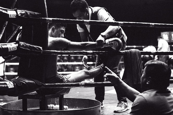 Boxing match corner view: boxer in ring getting attention from trainer, water bucket visible. Black and white photography.