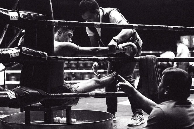 Boxing match corner view: boxer in ring getting attention from trainer, water bucket visible. Black and white photography.