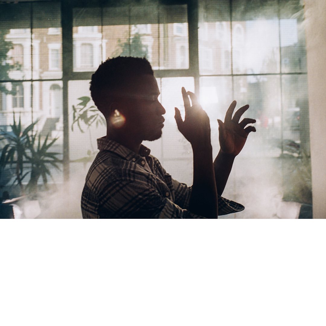 A dark-skinned adult male stands indoors near a window on a sunny day, holding his hands up to sunlight and wearing a plaid shirt.