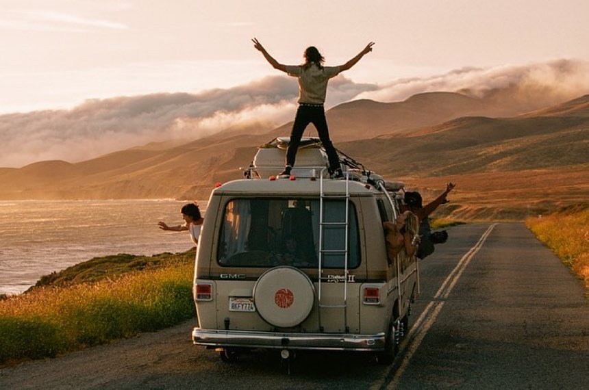 Group of friends on a road trip, one standing on top of a van with arms raised, with a coastal view and golden light at sunset.