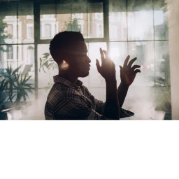 Silhouette of a Black man in a plaid shirt with hands raised, photographed against a large window by James Marcus Haney.