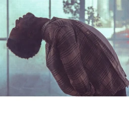 Man with beard arching backward wearing a plaid button-down shirt against a glass window background