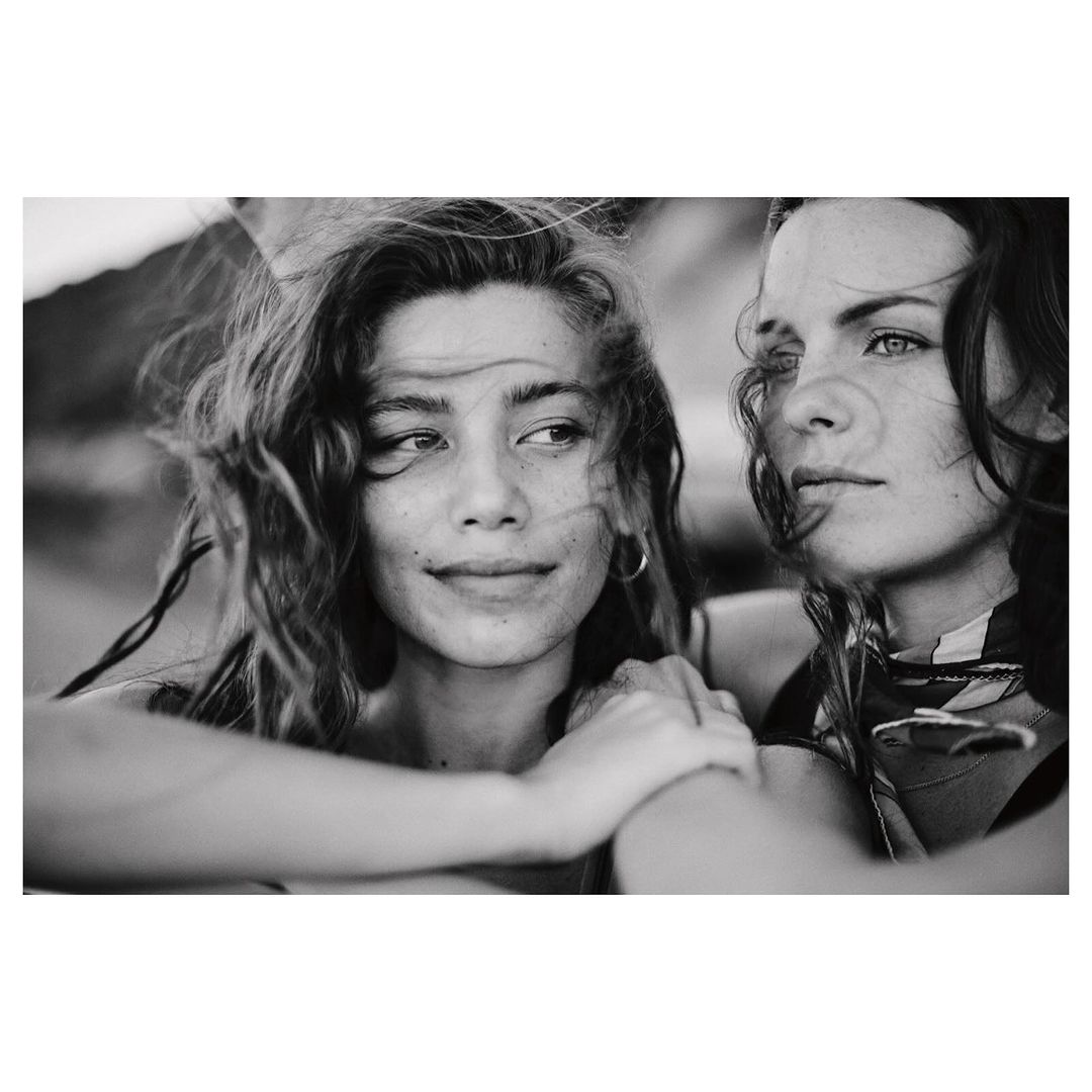 Black and white portrait of two women with tousled hair, close together, one smiling slightly, with an outdoor, natural light setting.
