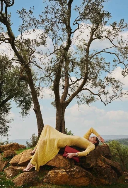 A woman in a yellow dress lies on rocks with a red clutch bag in an outdoor fashion shot, with a tree in the foreground against a blue sky.