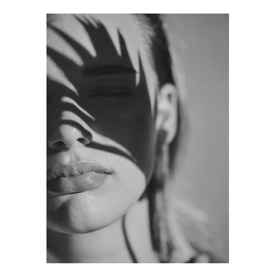 Monochromatic portrait of a woman with closed eyes and leaf shadows across her face in a minimalist composition, evoking natural light.
