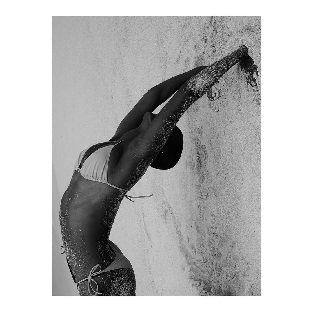 Woman in bikini stretches, reaching upwards with arms, covered in shimmering particles, set against a textured, neutral-toned wall.