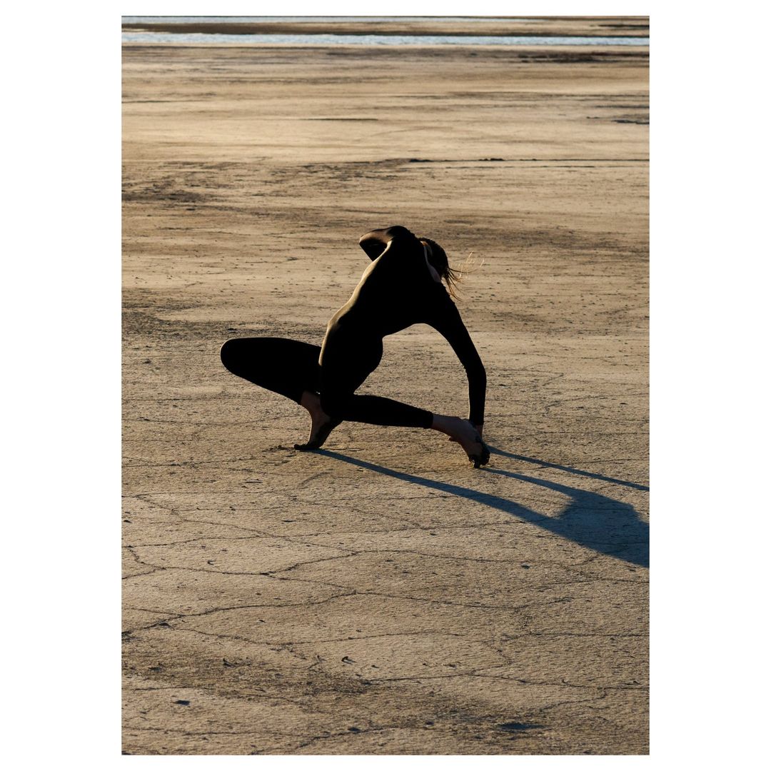 Silhouette of a person in a yoga pose on a cracked, sandy surface, with a blurred background, long shadow, and backlit effect.