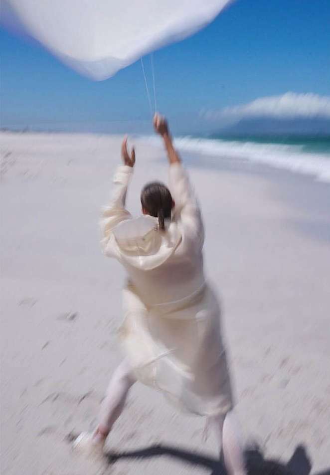A person in light-colored clothing walks on a beach, holding a white kite in the air, with the ocean and sky visible in the background.