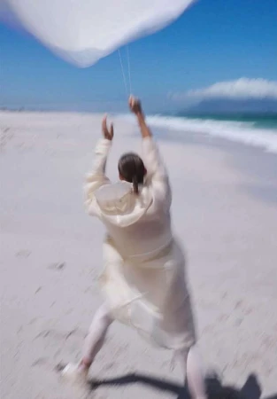 A person in light-colored clothing walks on a beach, holding a white kite in the air, with the ocean and sky visible in the background.