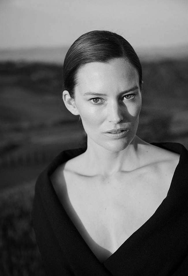 Black and white portrait of an adult female with dark clothing against an out-of-focus landscape, captured in natural, soft lighting.