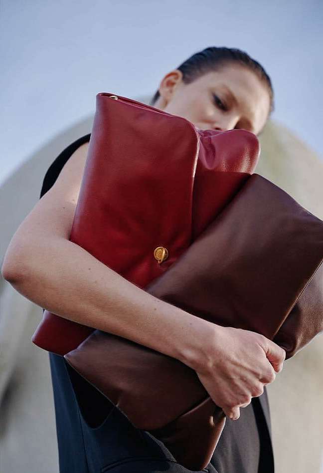 A woman holds a stack of three leather clutch bags in red and brown against a light blue sky and neutral architectural background.