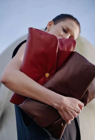 A woman holds a stack of three leather clutch bags in red and brown against a light blue sky and neutral architectural background.