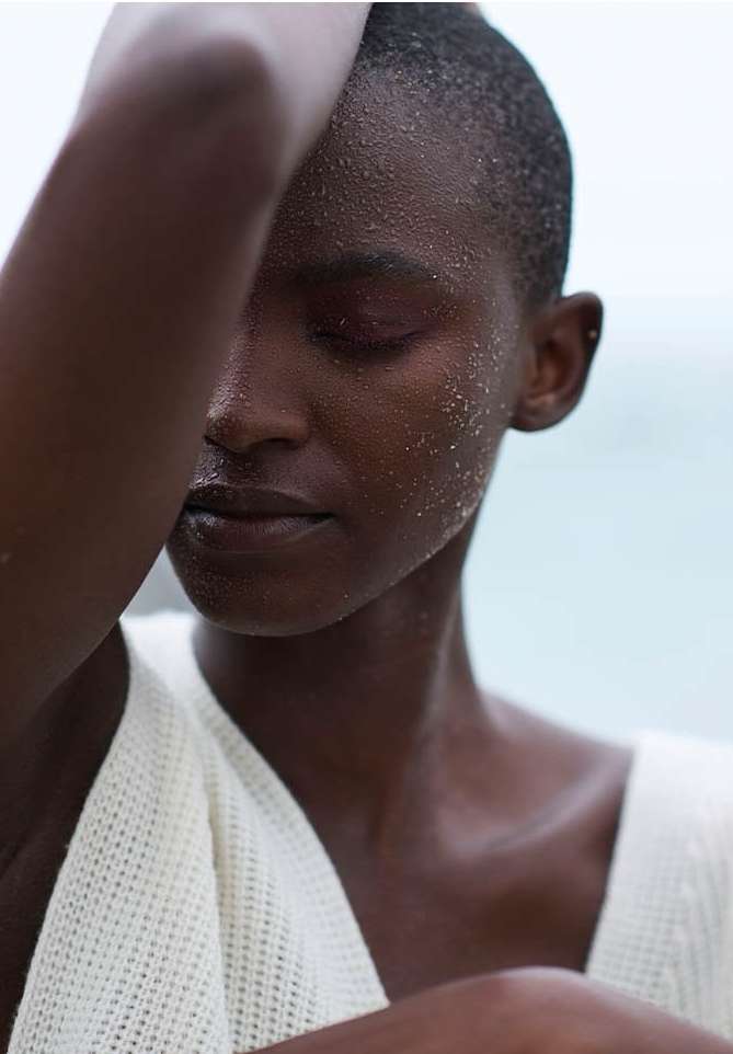 Close-up portrait of a Black female model with short hair, eyes closed, wearing a knitted top with a light blue background.