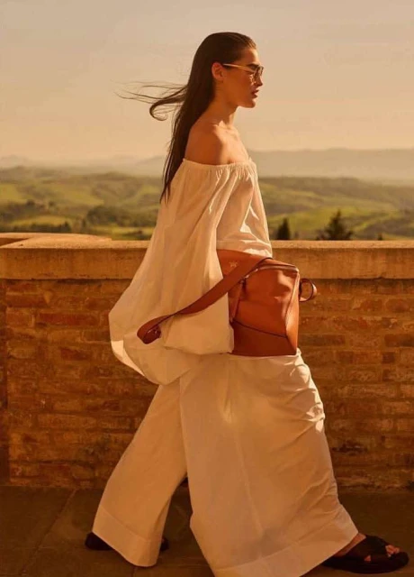 A woman in white off-the-shoulder top and pants walks with a brown bag against a warm landscape backdrop with rolling hills.