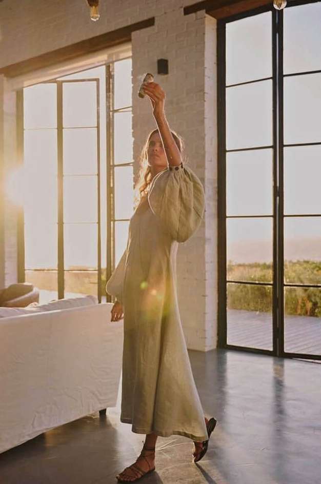 Adult female in a light-green dress poses with arms raised inside a sunlit home with large windows, concrete floors, and minimal decor.