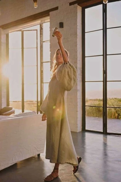 Adult female in a light-green dress poses with arms raised inside a sunlit home with large windows, concrete floors, and minimal decor.