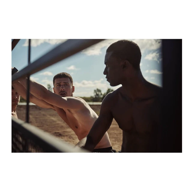 Two shirtless men stand outdoors near a metal fence; one is leaning on the fence, and the other stands beside it against a bright sky.