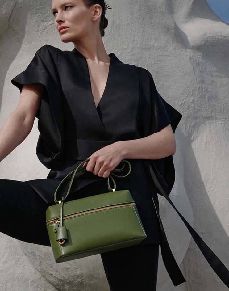 Fashion editorial shot of a woman holding an olive-green handbag, wearing a black wrap top and leggings against a textured wall.