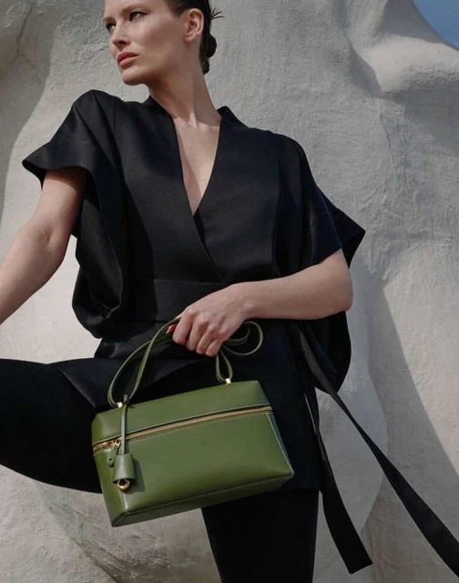 Fashion editorial shot of a woman holding an olive-green handbag, wearing a black wrap top and leggings against a textured wall.