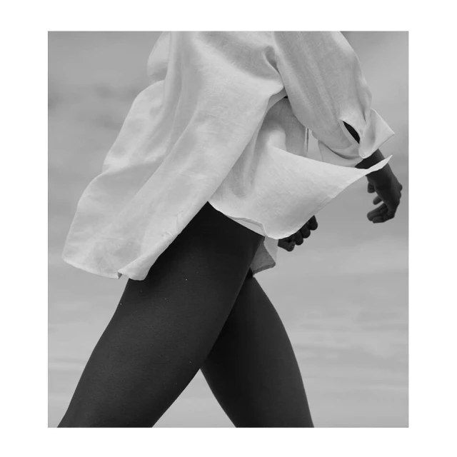 Monochromatic fashion shot of a woman in a billowing white linen shirt, walking, with her dark skin contrasting against the light fabric.