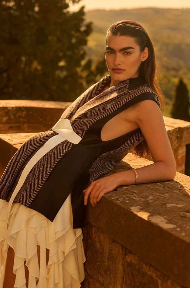A woman poses in a sleeveless top and white skirt against a Tuscan landscape at golden hour, with a stone wall in the foreground.