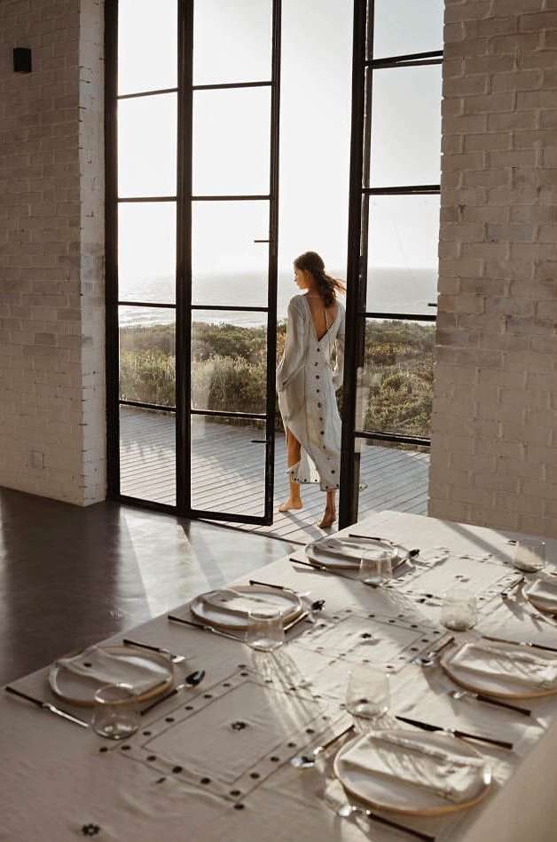 Back view of woman in flowing dress standing in open doorway looking at ocean; place settings visible on dining table.
