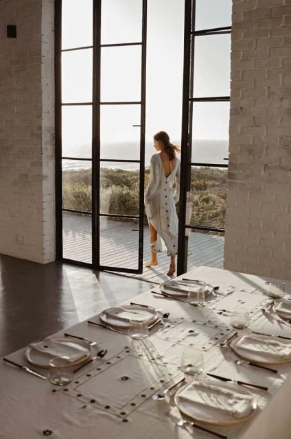 Back view of woman in flowing dress standing in open doorway looking at ocean; place settings visible on dining table.