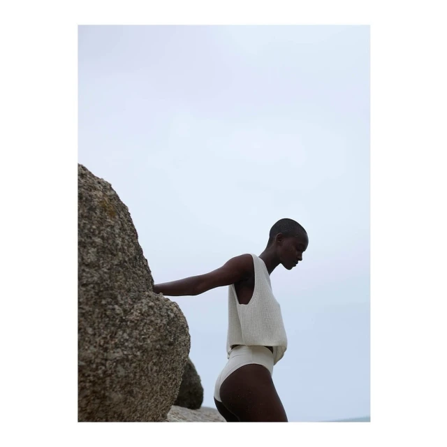 An adult female poses, leaning on a rock formation near the ocean, wearing a matching cream-colored knitted crop top and high-waisted bottoms.
