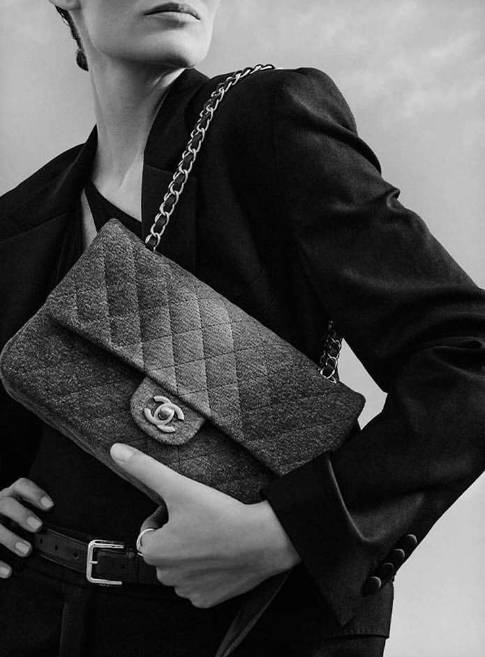 A woman in a dark blazer poses with a Chanel flap bag featuring a chain strap, highlighting a fashion-forward aesthetic.