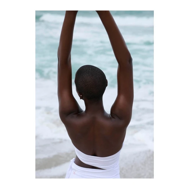 A black woman stands on a sandy beach with her arms raised, wearing a white wrap. The ocean is visible in the background.