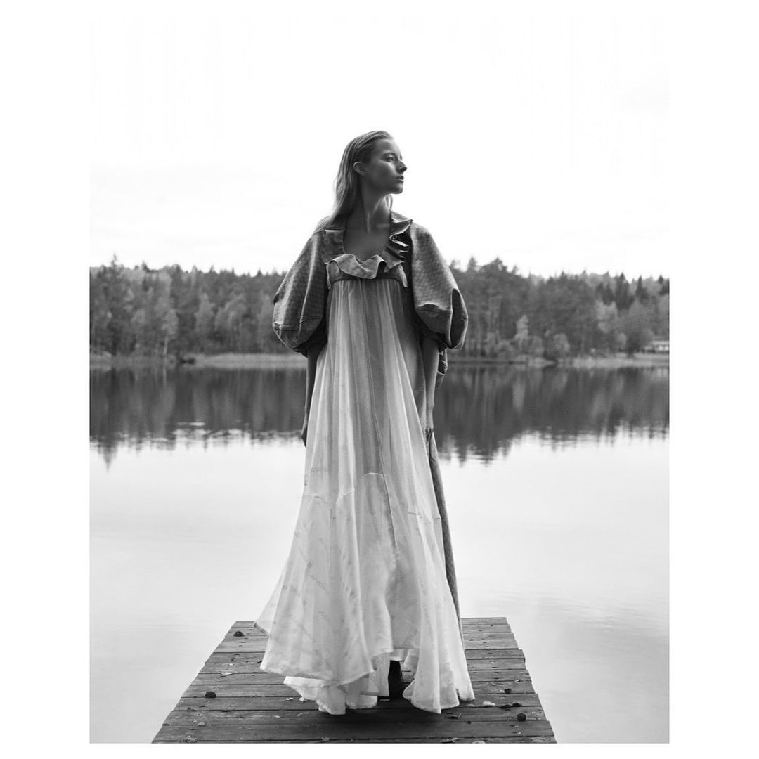 Woman in a white dress and grey jacket standing on a wooden dock by a tranquil lake with a forest backdrop in a black and white portrait.