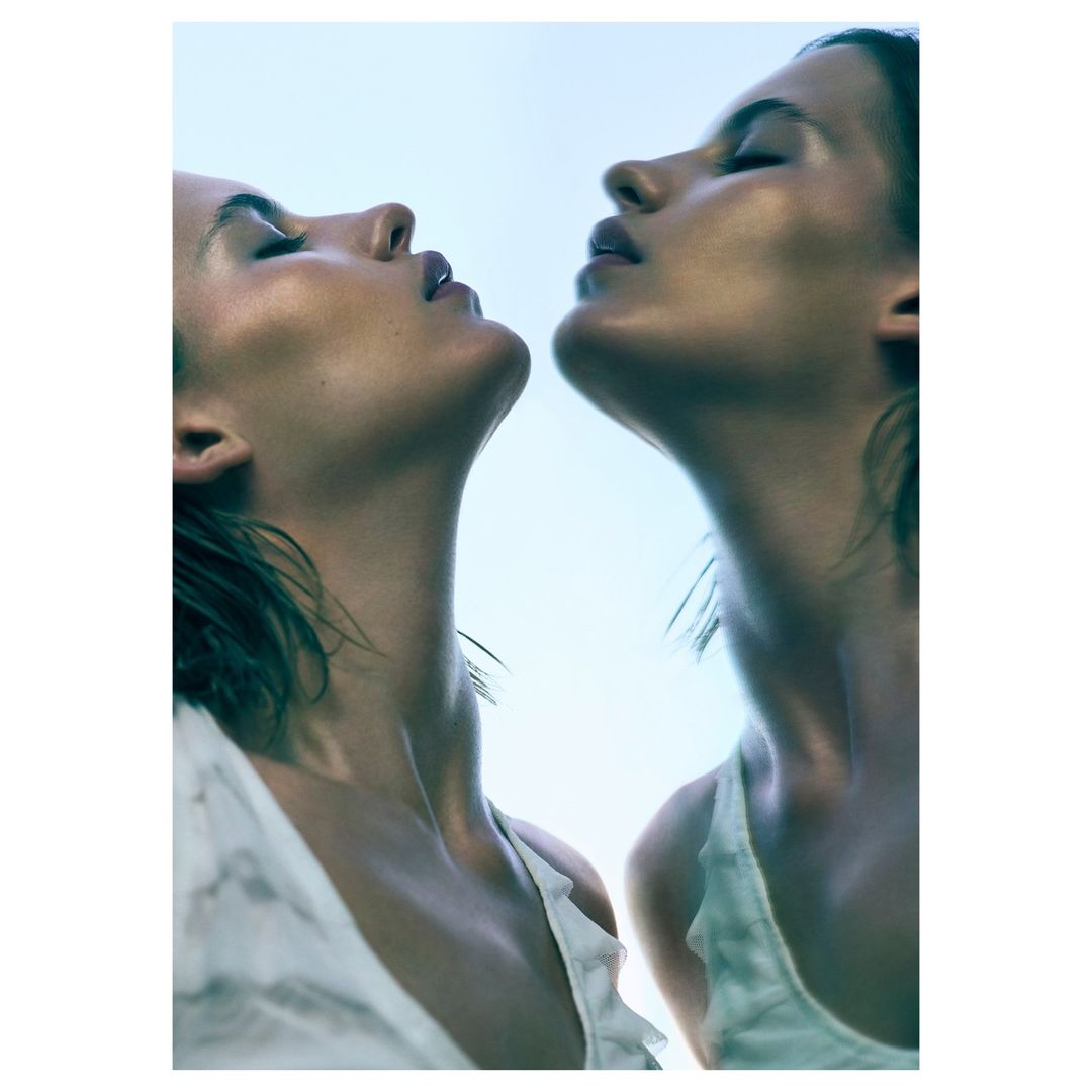 Two adult women pose together in a fashion headshot, looking up with neutral expressions in a photo studio with soft lighting.