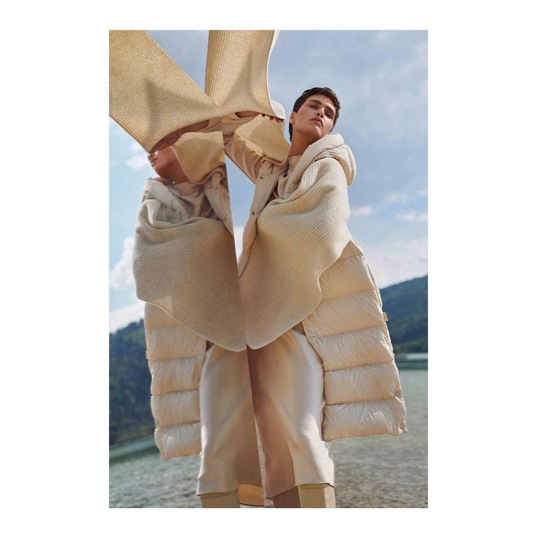 Fashion shot of a woman in a padded coat and loose pants stands outdoors against a backdrop of water and mountains with a sky view.