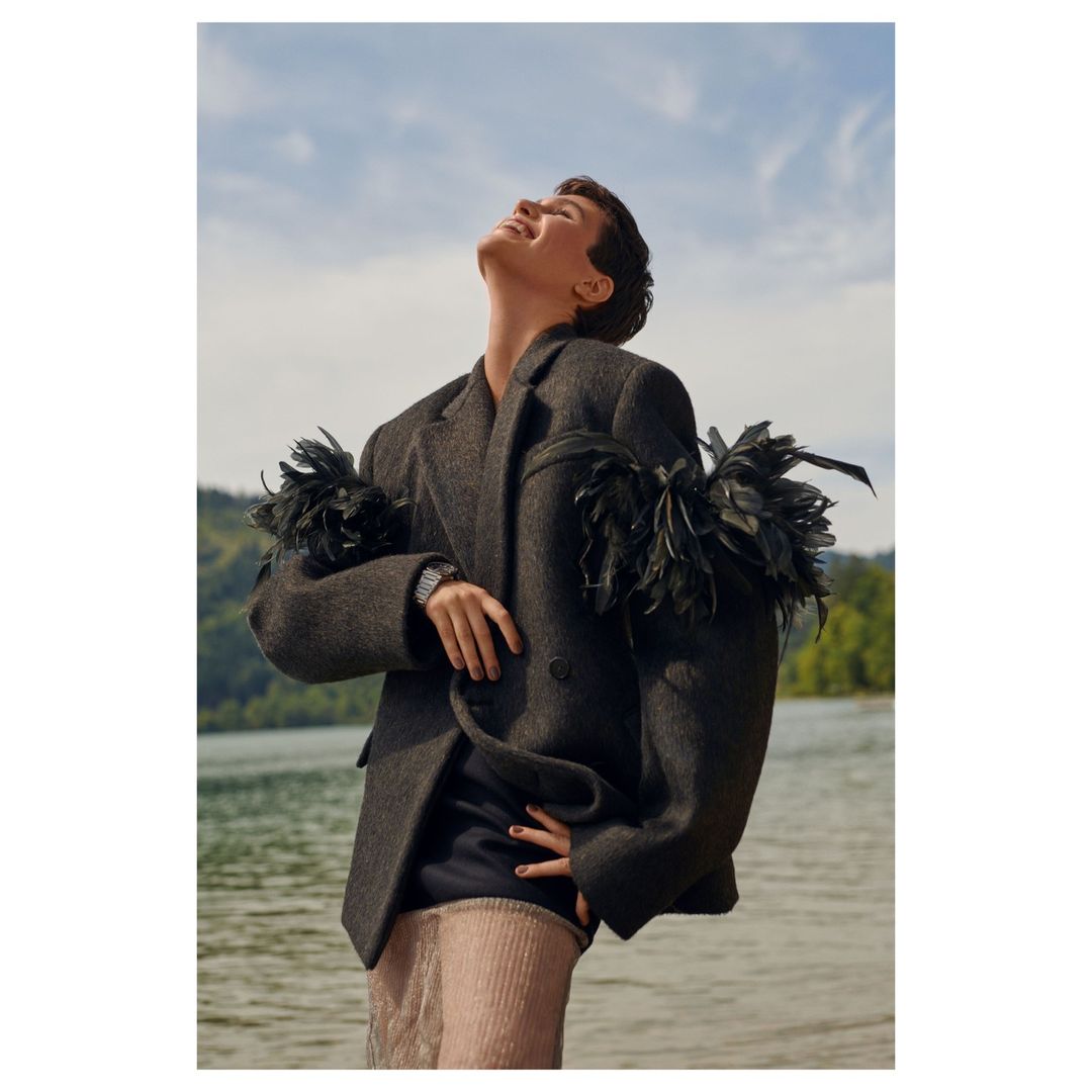 Outdoor portrait of a person in a feathered jacket, basking in natural light by a lake surrounded by trees, under a bright blue sky.