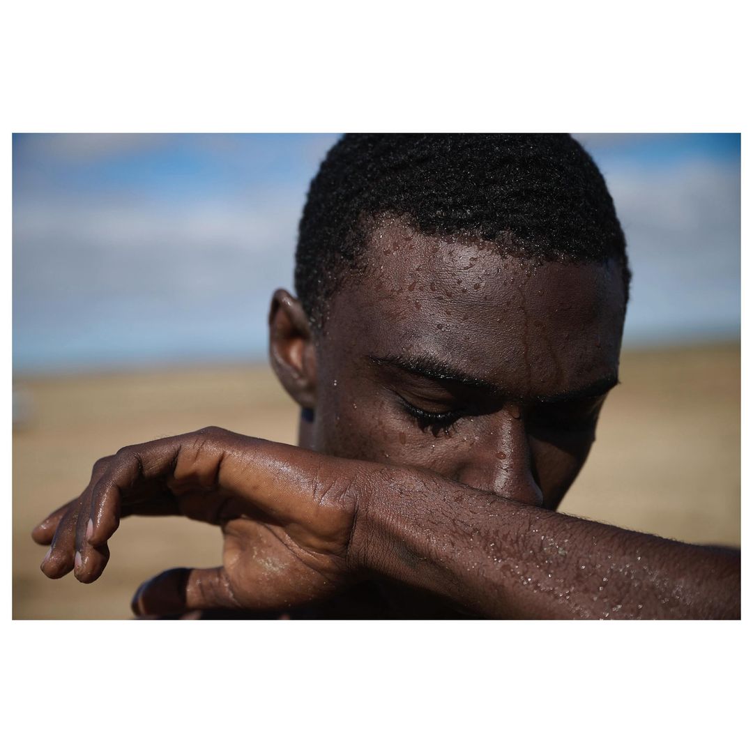 Close-up portrait of a sweaty Black male model outdoors on a sandy surface with a blue sky; shows texture and lighting details.
