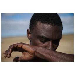 Close-up portrait of a young Black man wiping sweat from his face against an outdoor landscape background.