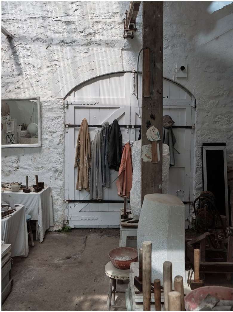 Brightly lit artist's studio featuring finished sculptures, tools, and work surfaces against a textured white brick wall and door.