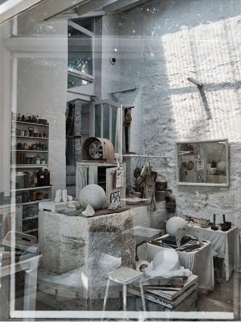 Artist's studio featuring sculptures, tools, and art pieces arranged on tables and shelves in a bright, creative workspace.